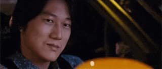 Han de Fast & Furious : Tokyo Drift est assis dans une voiture dans le siège passager, regarde devant soi avec un air un peu joueur et il sourit, hoche la tête légèrement puis la voiture démarre et il disparaît du plan de caméra aussitôt.