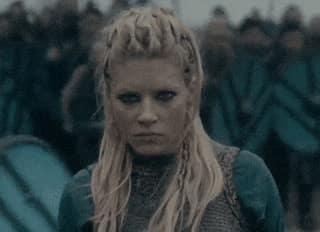Lagertha de la série Vikings en plein champ de bataille, vue de près, avec un air stoïque, pendant que des guerriers avancent à ses côtés.