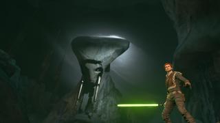 Cal se tient debout devant une énorme statue d'un personnage alien, dans une caverne seulement éclairée par un trou dans le plafond au-dessus de la statue.
