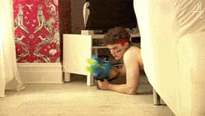 Un jeune homme avec deux bandes peintes sous les yeux et un badana rouge autour de la tête tient un pistolet à eau avec de vives couleurs et rampe au sol dans un salon moderne, tout en ayant l'air très sérieux.