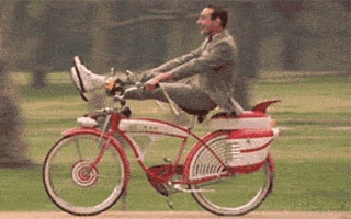 Un homme sur un vélo rouge vintage avance rapidement dans un parc, les pieds posés sur le guidon, tout en équilibre.